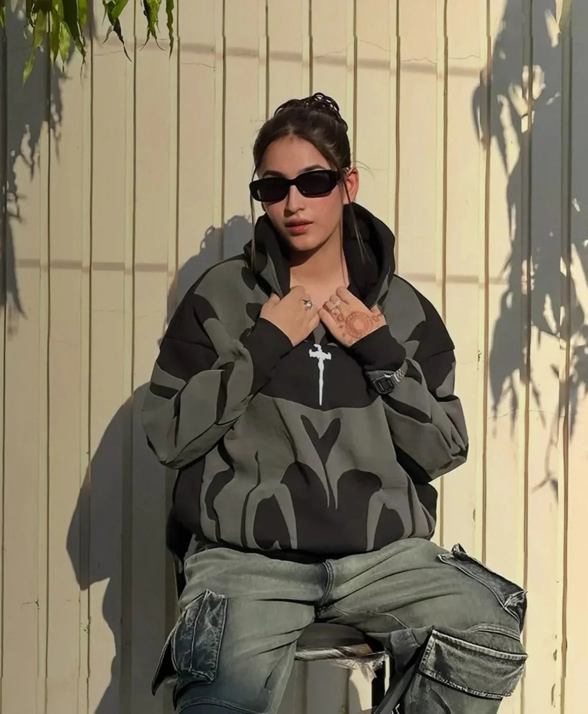 Person wearing a camouflage hoodie and sunglasses sitting against a light-colored wall.