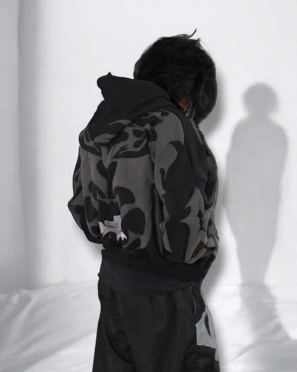 Person wearing a black and gray patterned hoodie with a logo, standing against a white wall.